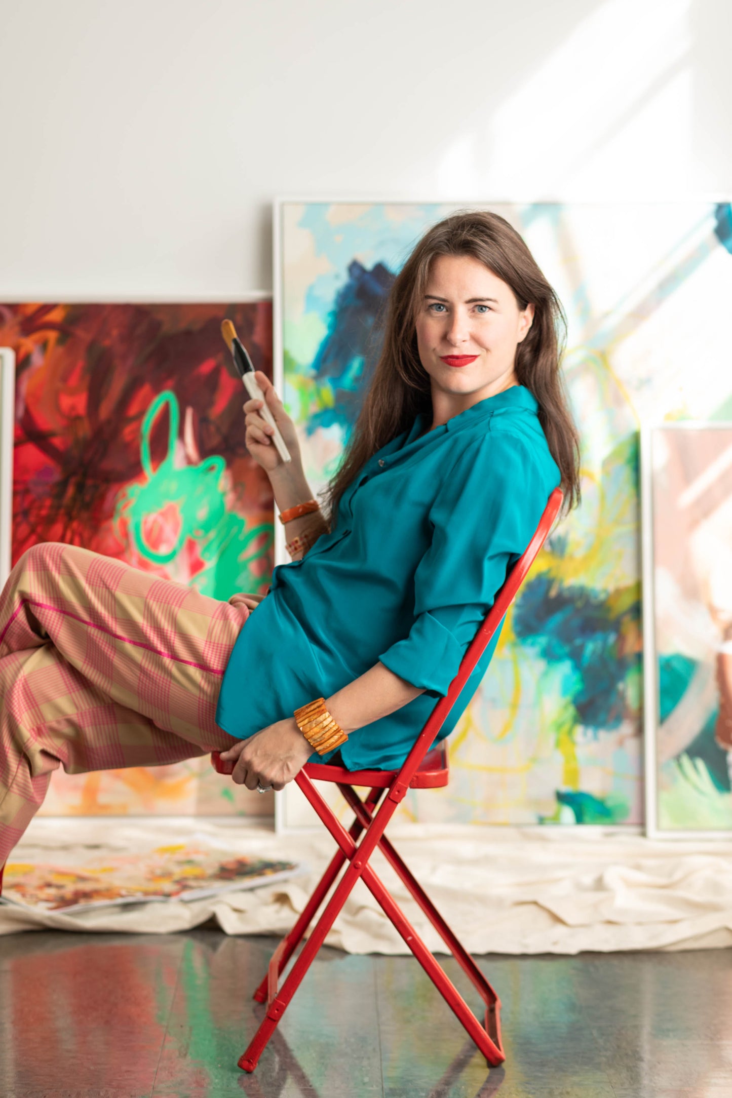 New England artist Megan Carty sitting on a red stool in front of colorful abstract paintings.