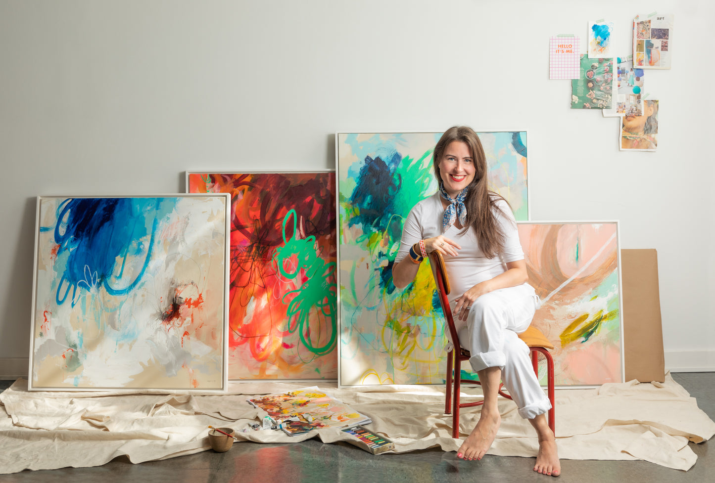 New England artist Megan Carty sitting on a chair surrounded by her energetic colorful intuitive abstract paintings in a studio setting.