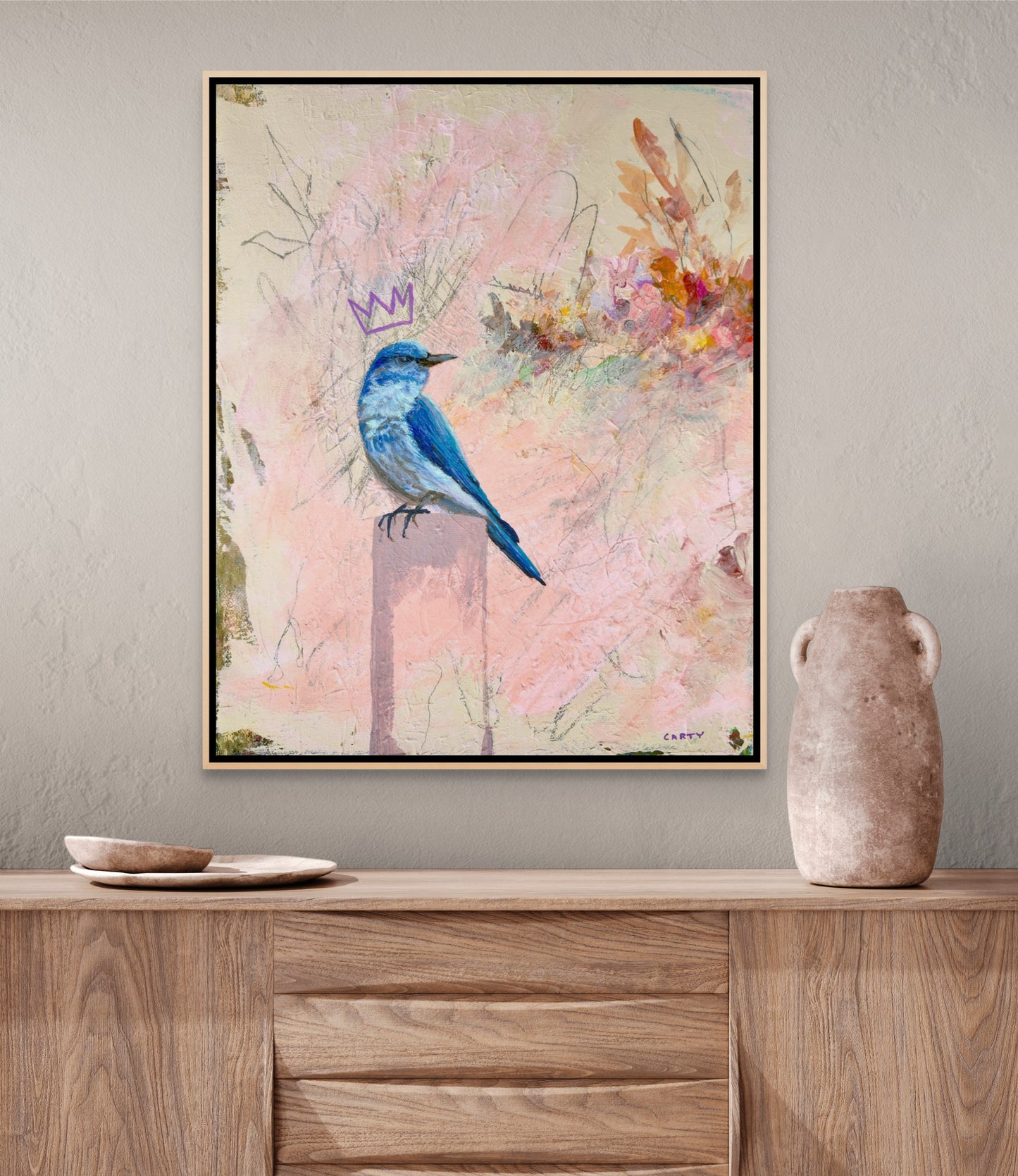 Framed artwork of a blue bird with a crown painted by Groton, MA artist Megan Carty on a wall above a wooden cabinet.