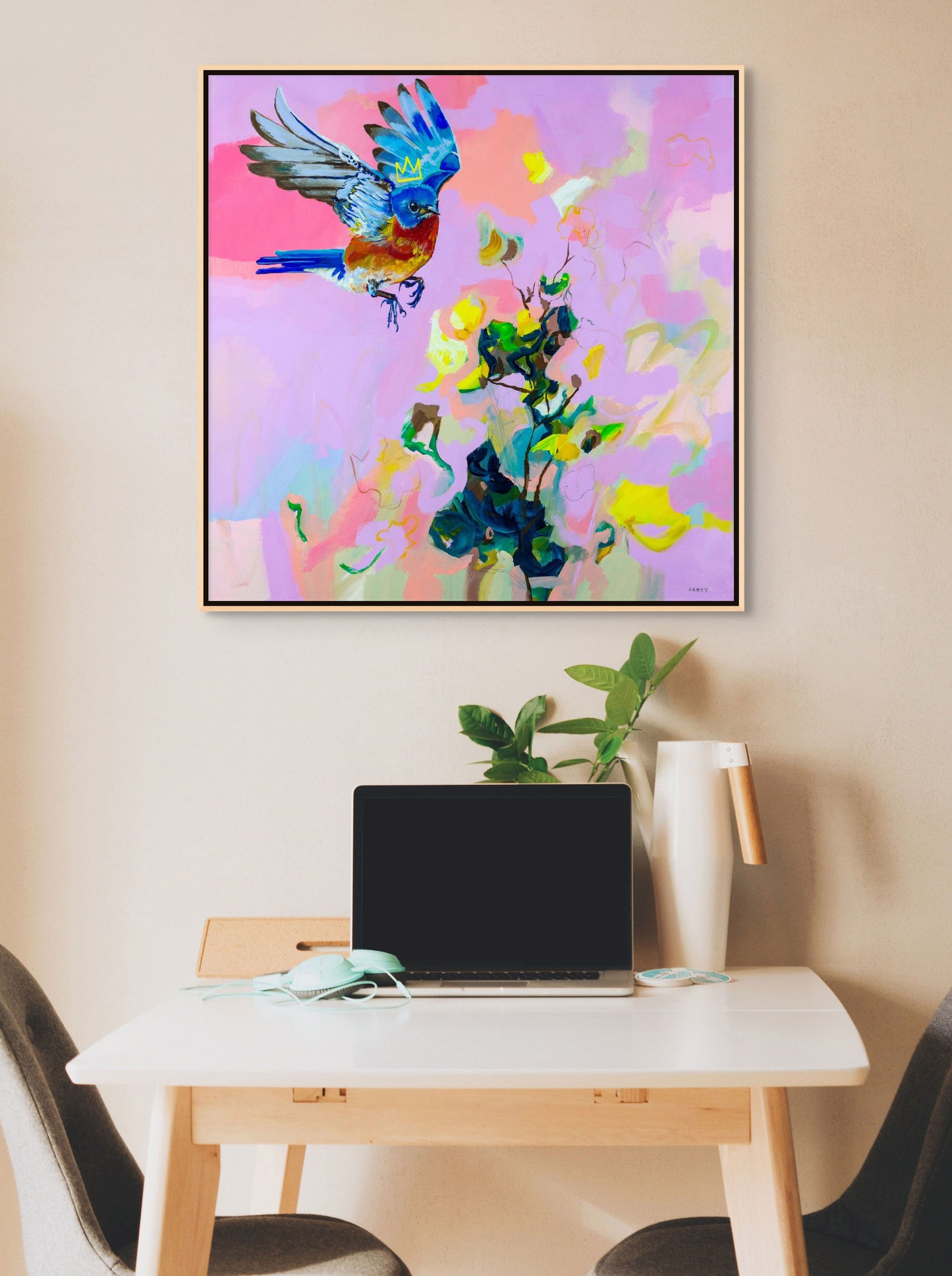 Framed abstract floral artwork with a bluebird and flowers painted by Boston area artist Megan Carty. hanging over a small table with a laptop and modern chairs.