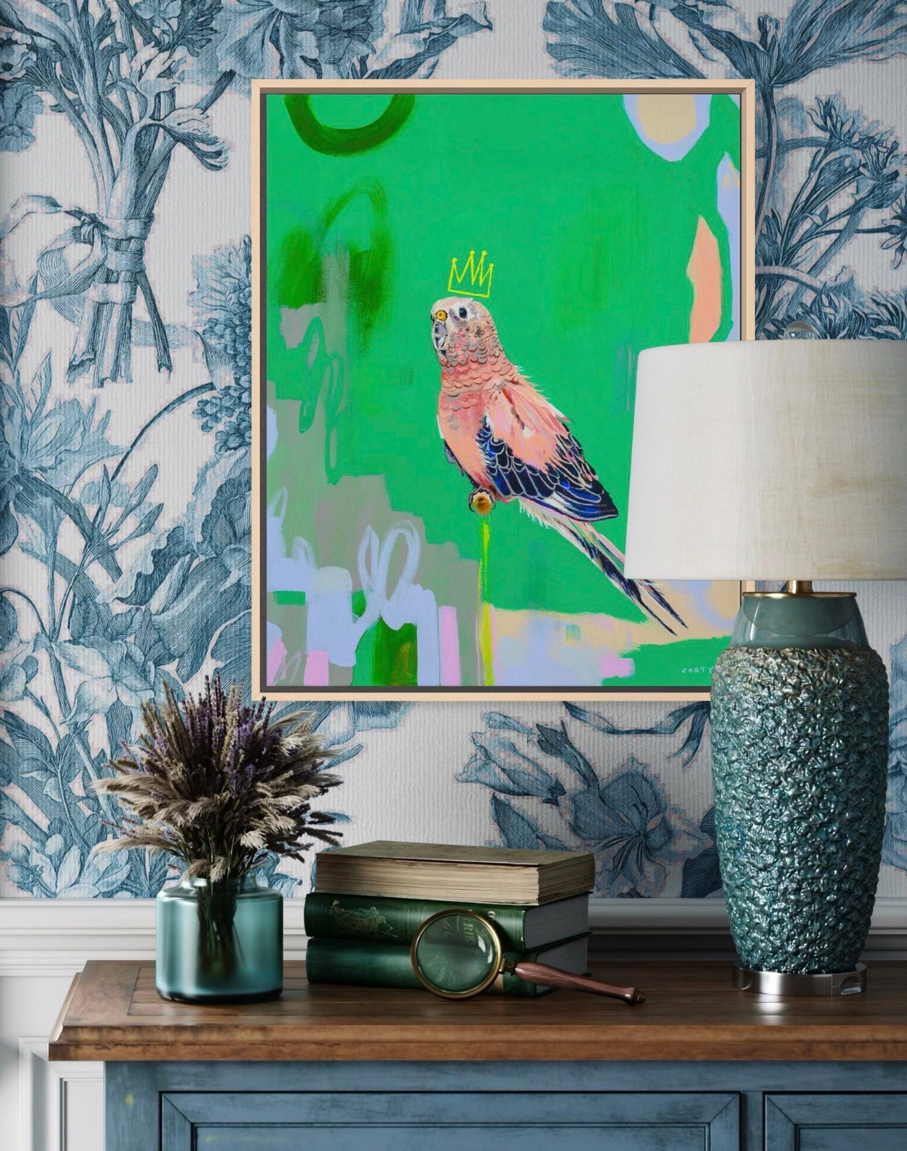 Decorative setting with a colorful parrot painting by New England artist Megan Carty, lamp, books, and plant on a wooden surface against floral wallpaper.